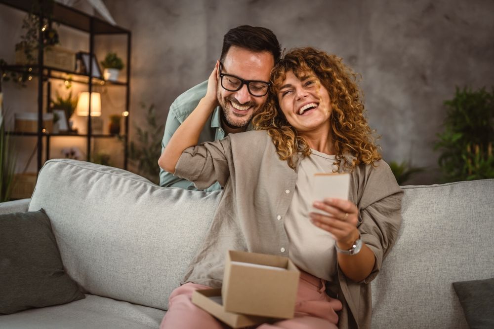 Man behind woman, looking at gift. Woman smiles, holding a box. Living room setting.