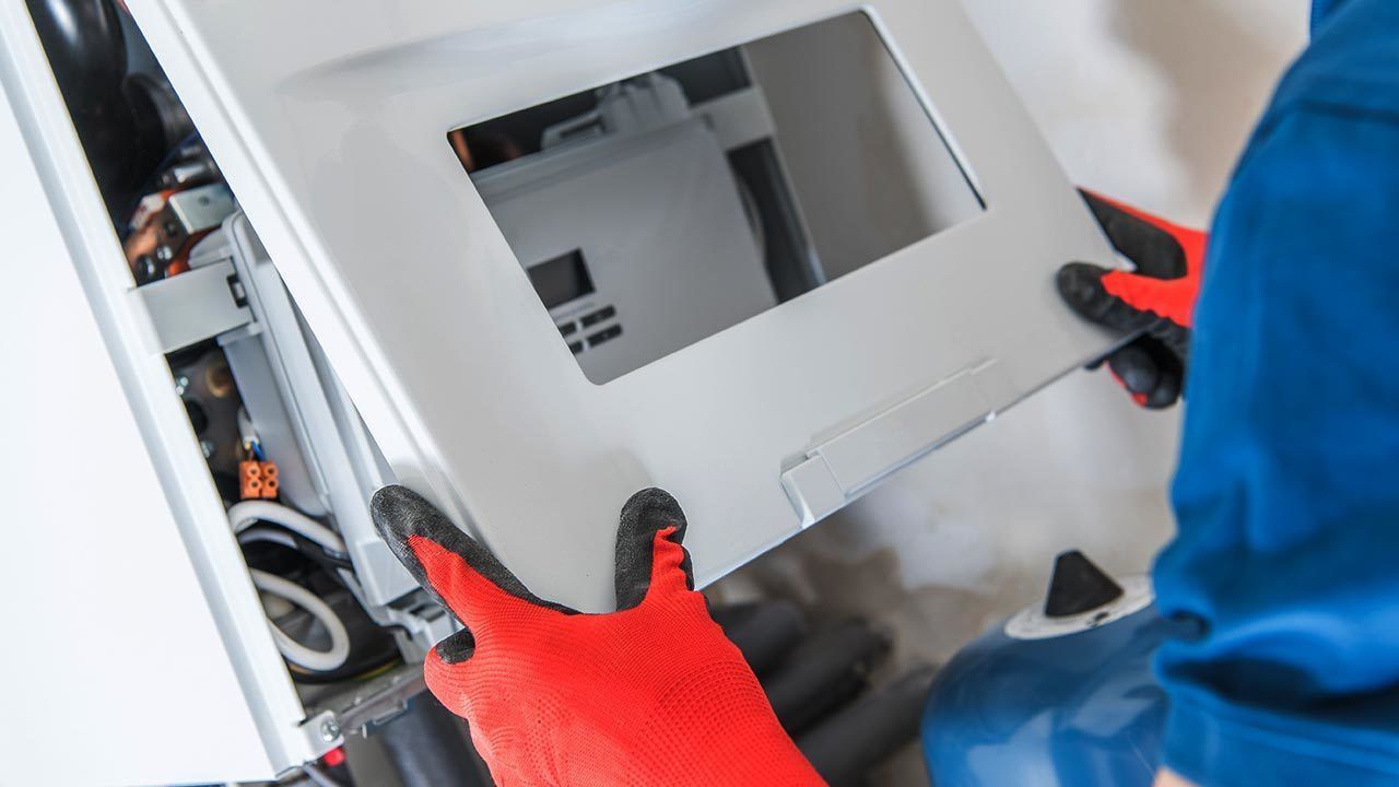 A person in red gloves and blue shirt working on a furnace, holding a metal panel.