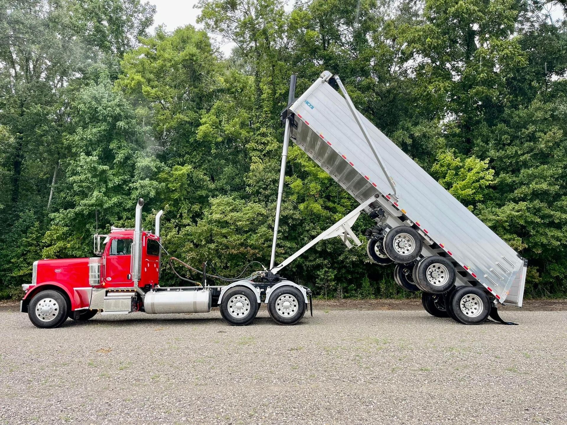 Red semi-truck with tilted dump trailer. Trailer bed is raised, releasing its contents. Trees in background.