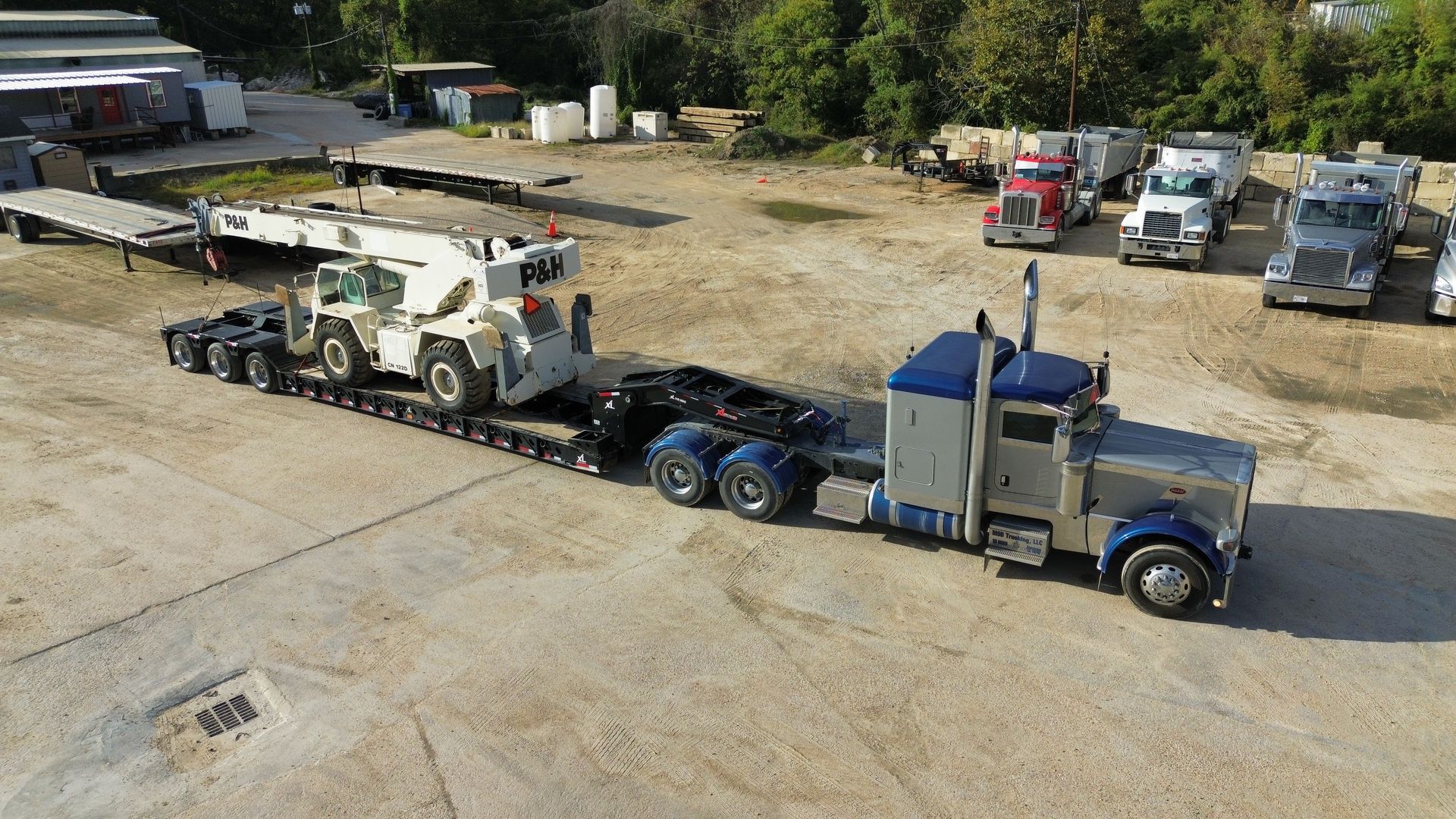 Heavy-duty truck hauling a large crane on a flatbed trailer in a gravel yard, with other trucks in the background.