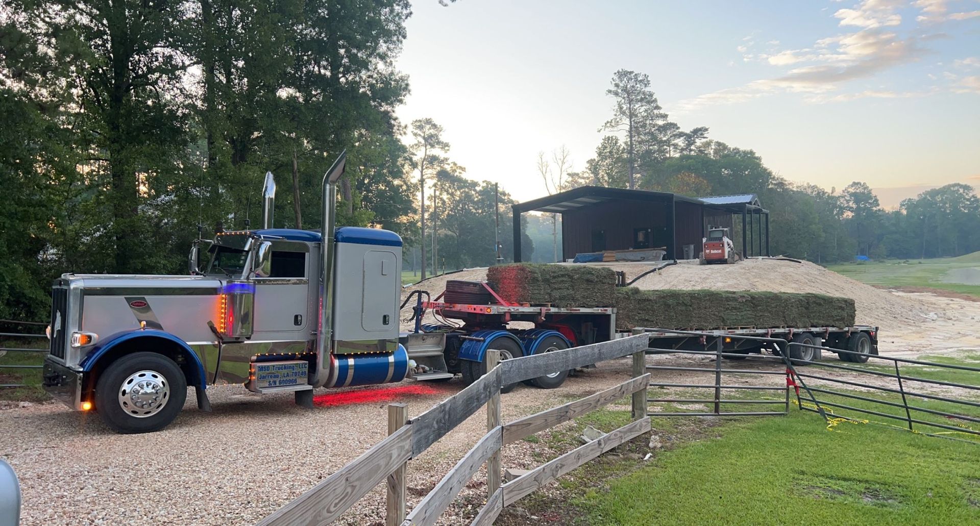 Semi-truck hauling hay bales near a barn. Blue truck with chrome details, brown fence in foreground.