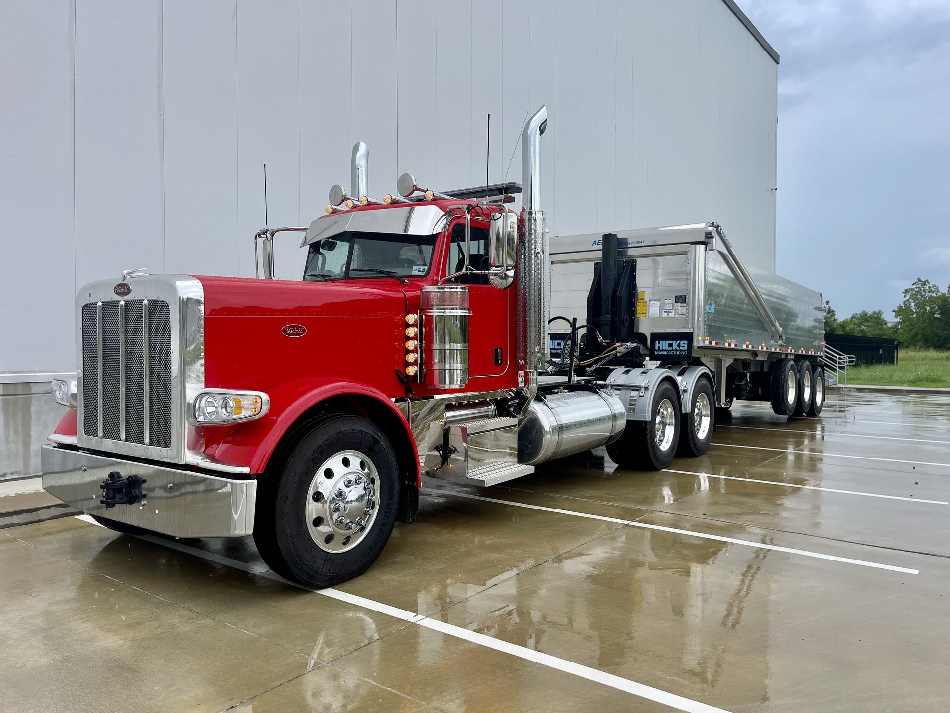 Red Peterbilt semi-truck with chrome details and a silver trailer parked on wet pavement.