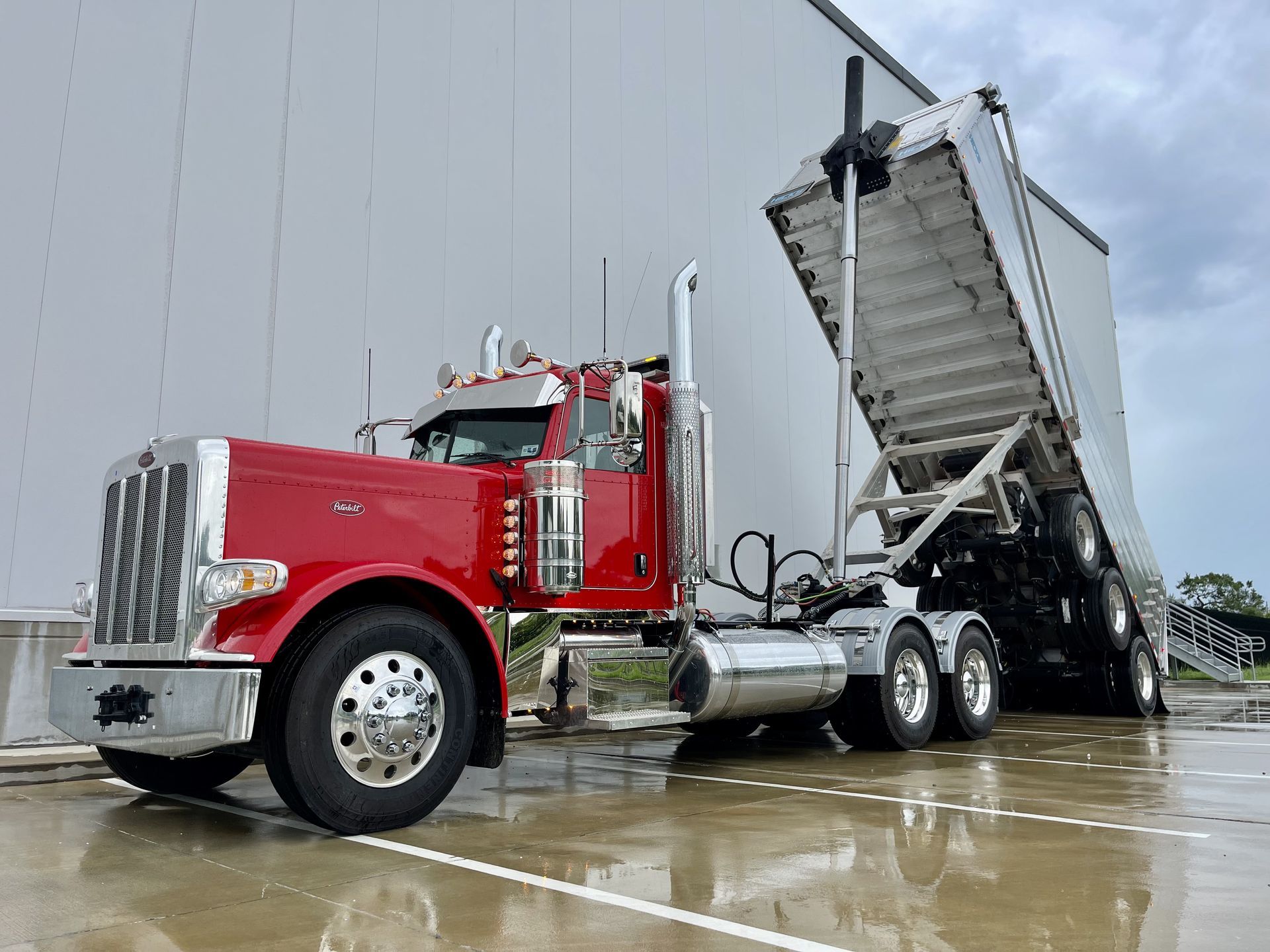 Red Peterbilt semi-truck with tilted dump trailer. Dumping cargo. Outdoors, against a gray building.