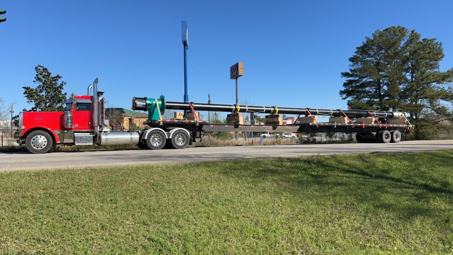 Red semi-truck hauling long metal beams on a flatbed trailer, driving along a road on a sunny day.