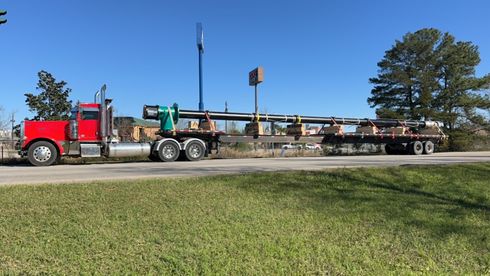 Red semi-truck hauling long metal beams on a flatbed trailer, driving along a road on a sunny day.
