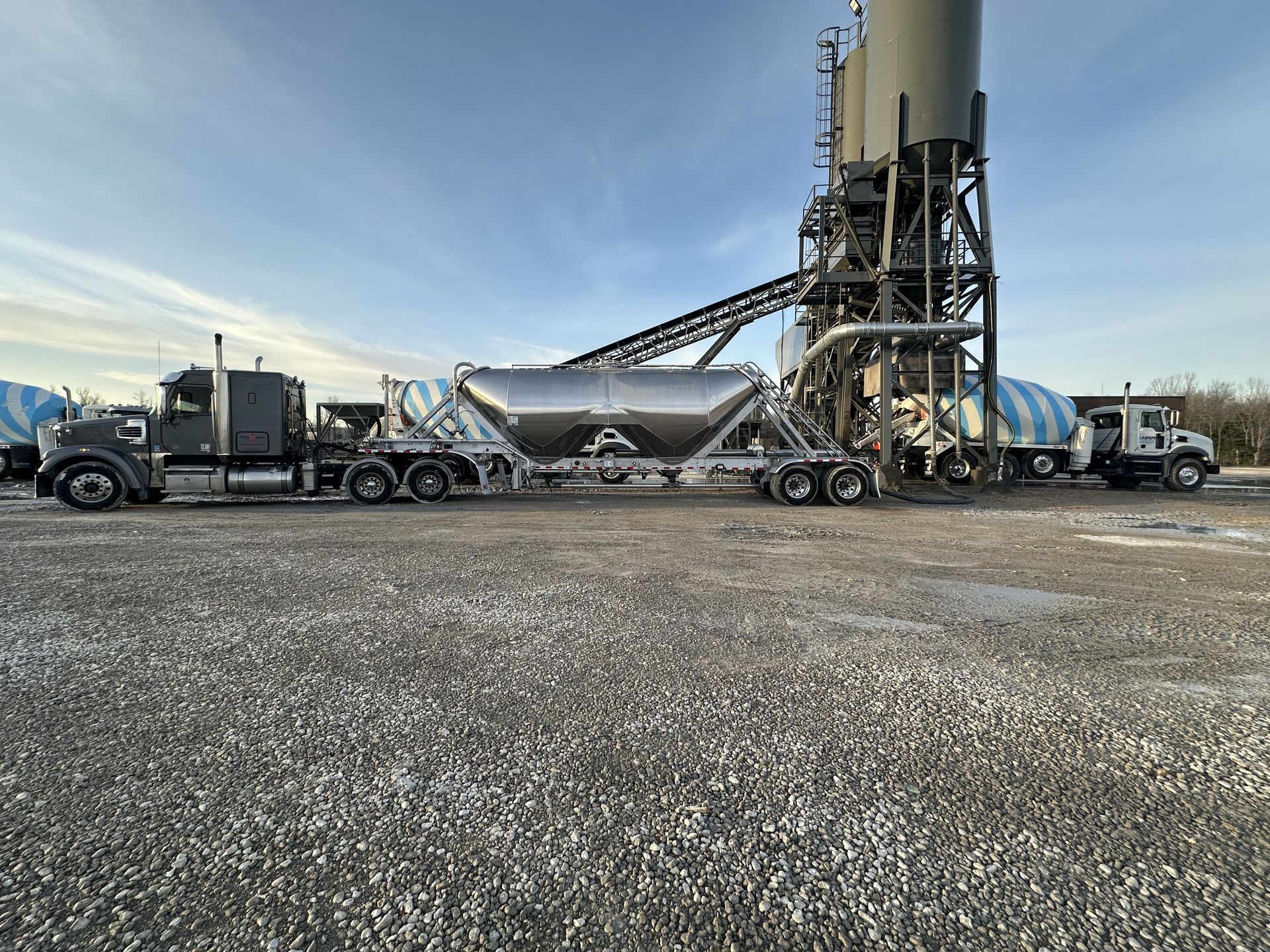 Semi-truck with tanker trailer at a concrete plant, loading material.