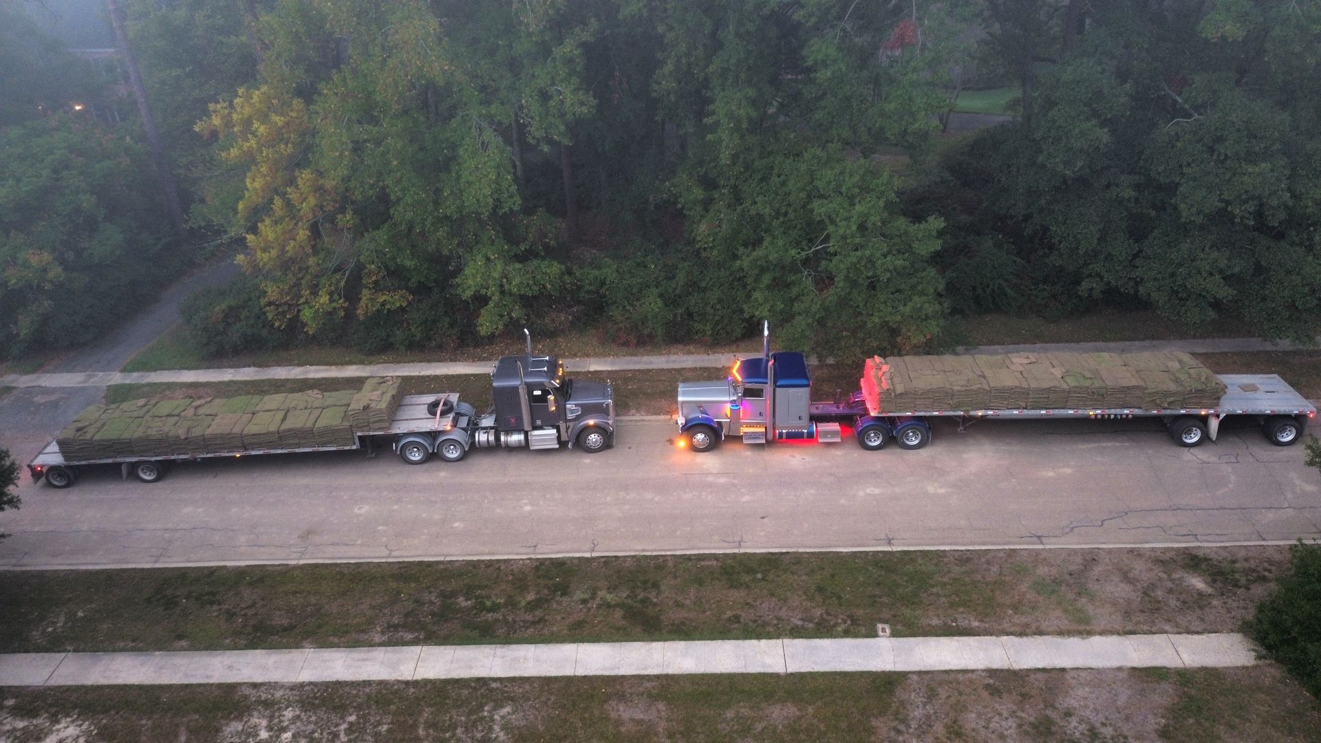 Two semi-trucks with trailers loaded with hay on a road, trees in background, overcast day.