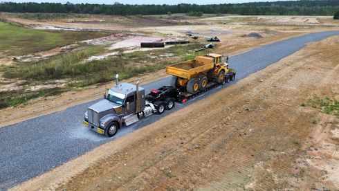 Truck hauling a large yellow dump truck on a gravel road, construction site in the background.
