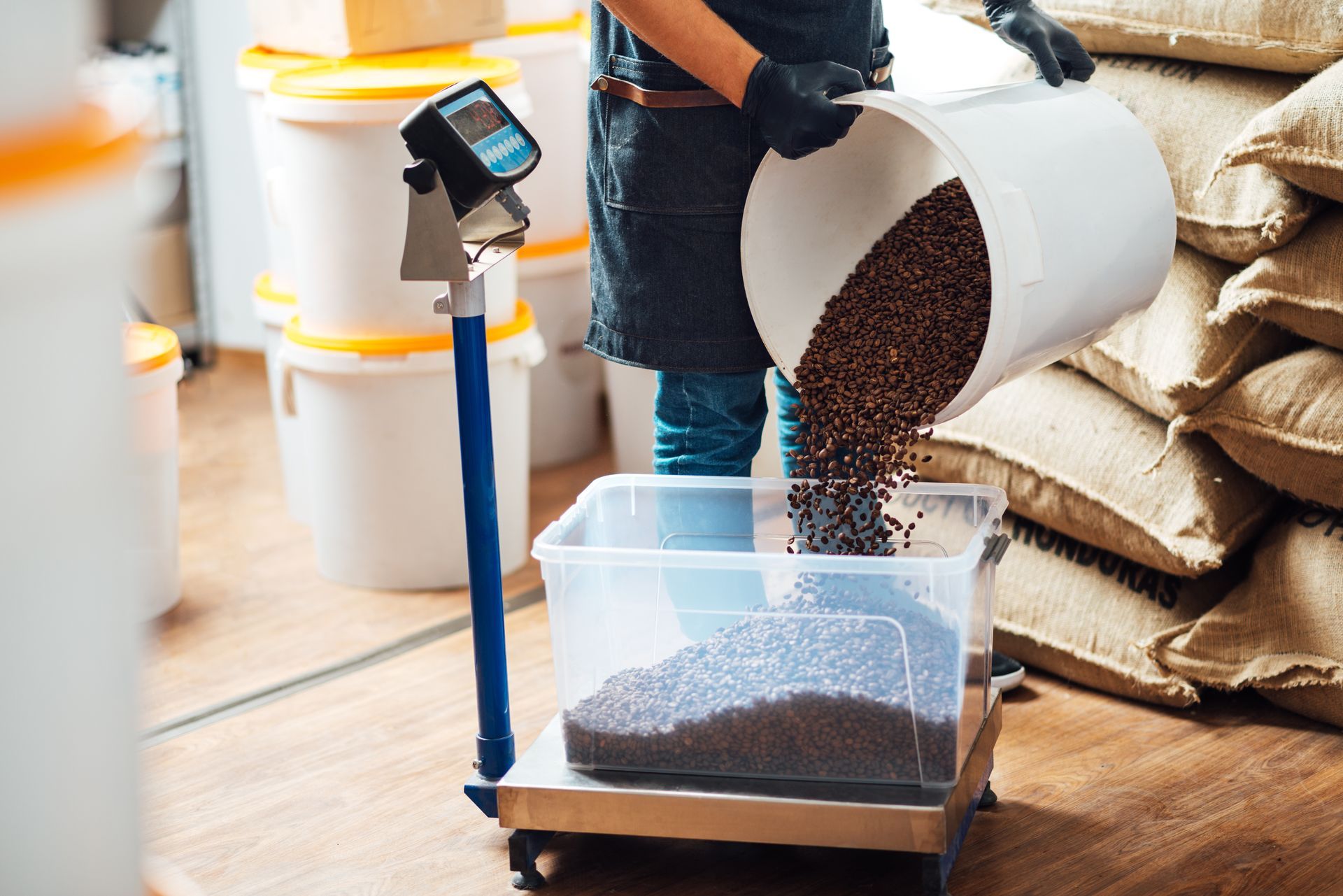 A person pouring coffee beans from a white bucket into a clear container on a digital scale in a warehouse.