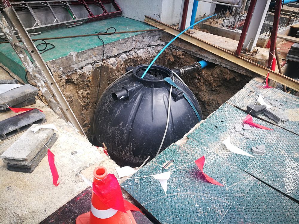 Black spherical tank in a construction pit, connected to blue and black pipes, surrounded by soil and construction materials.