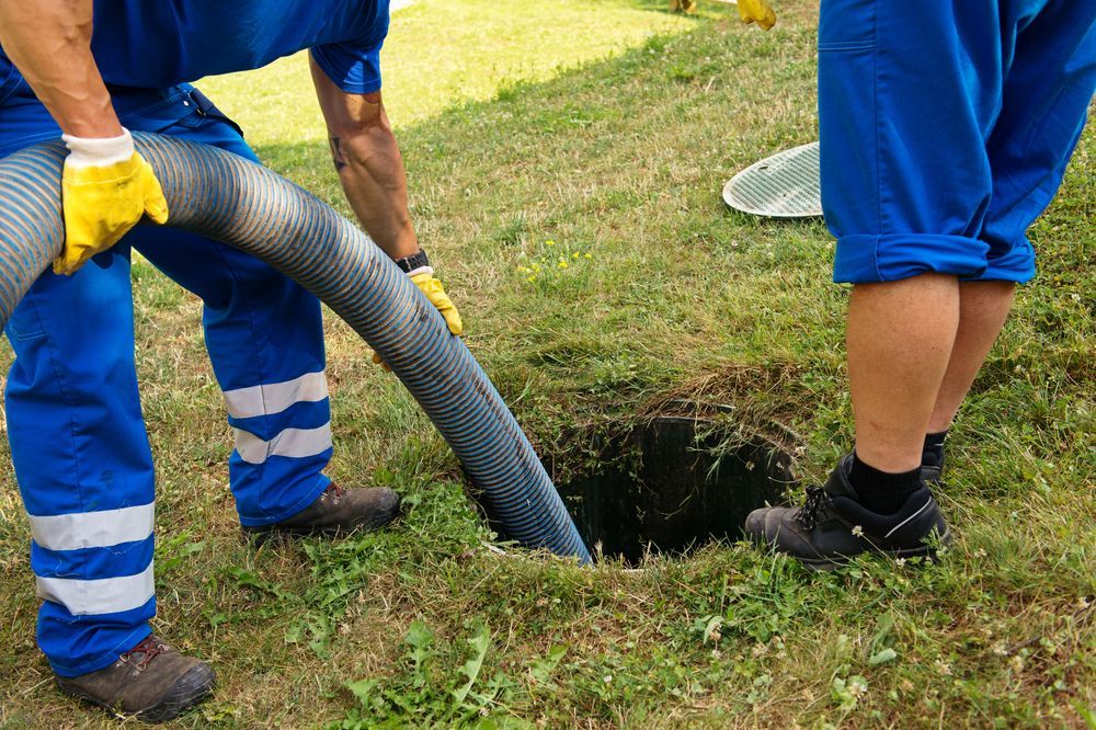 Two workers pumping a septic tank. One holds hose, other stands near the open tank.