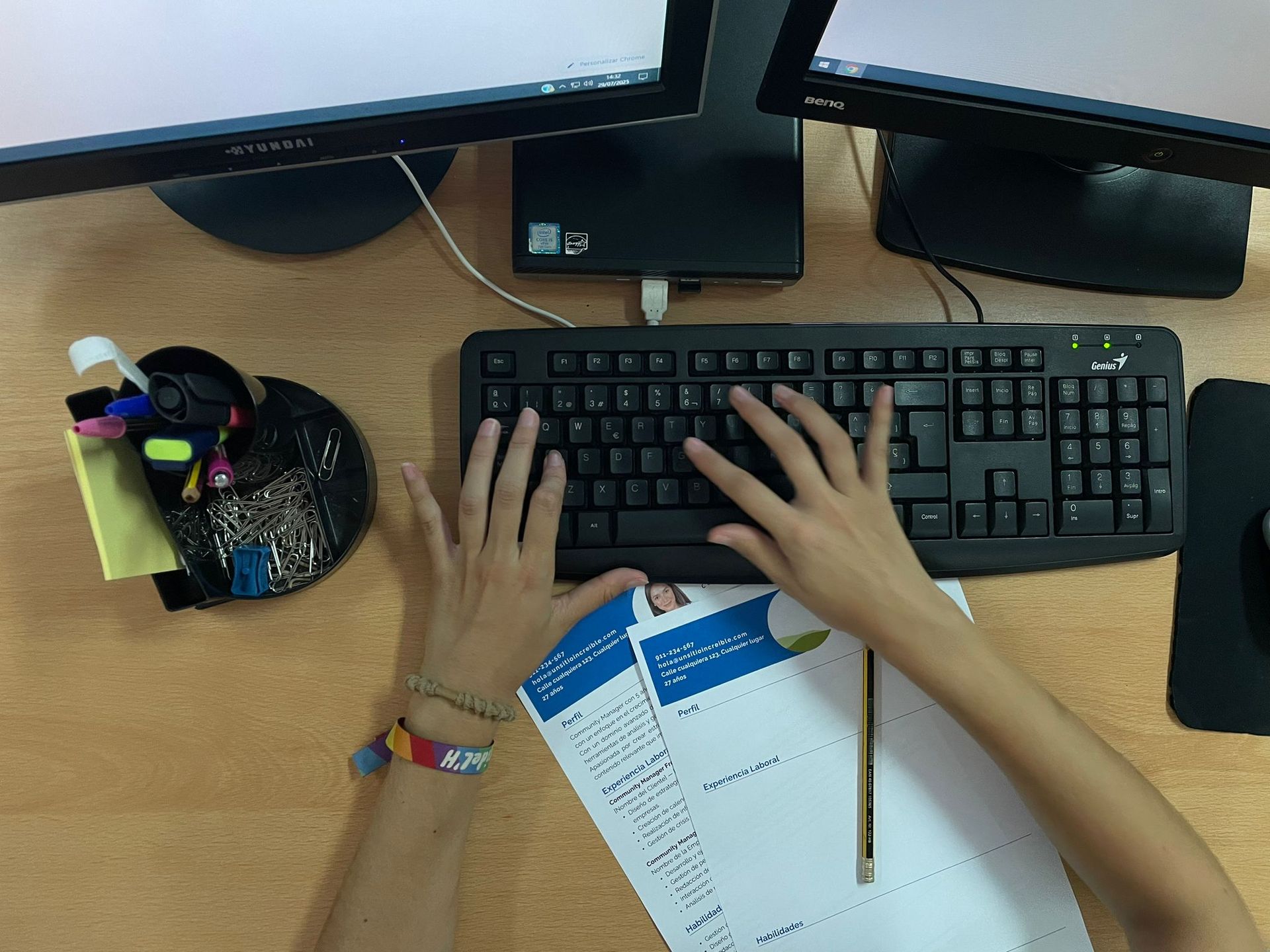 Mans escrivint en un teclat, papers i llapis visibles sobre un escriptori amb tres monitors.