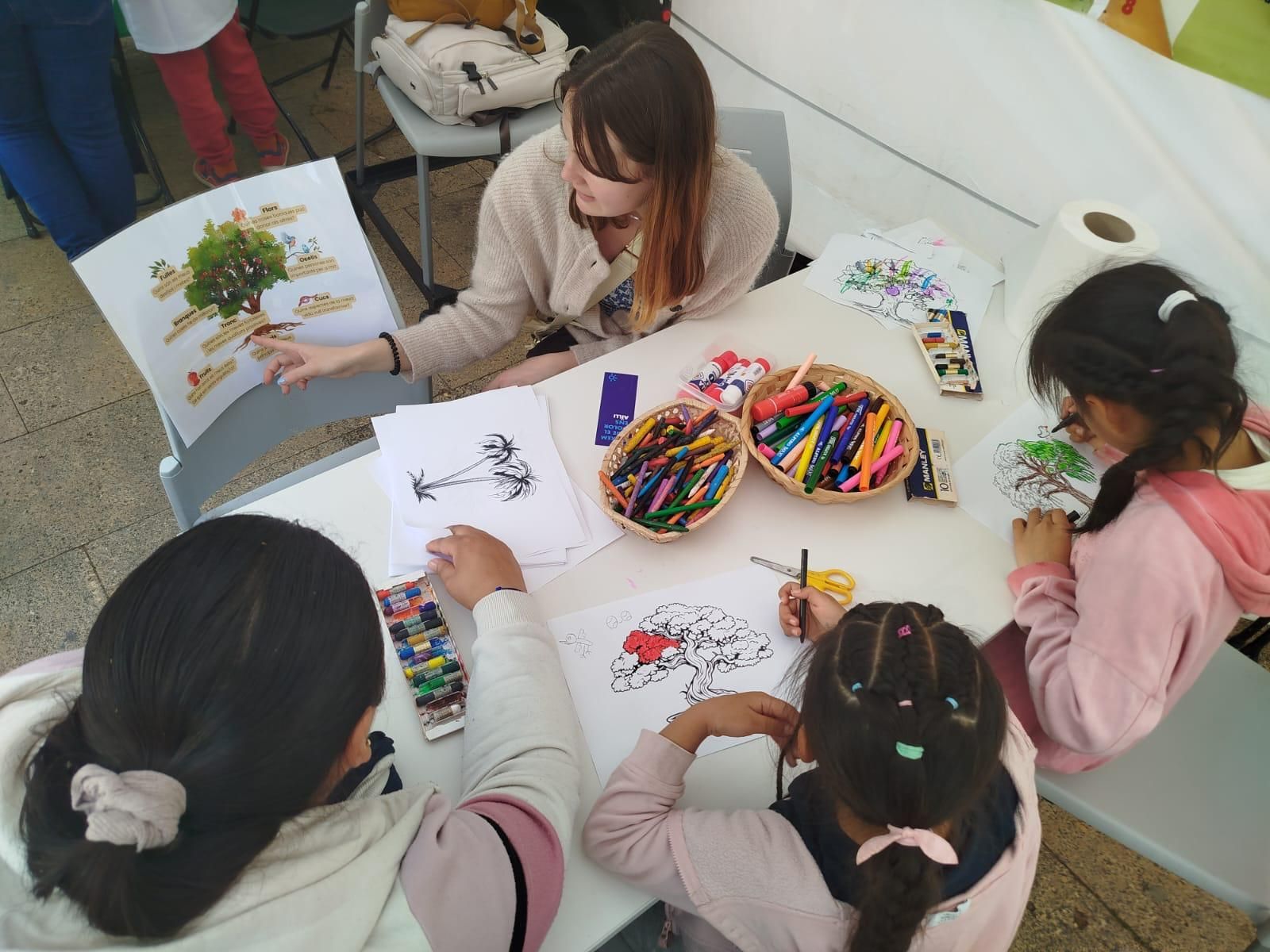 Una dona llegeix a tres noies assegudes a una taula amb llapis de colors i pàgines per pintar, probablement una activitat artística.