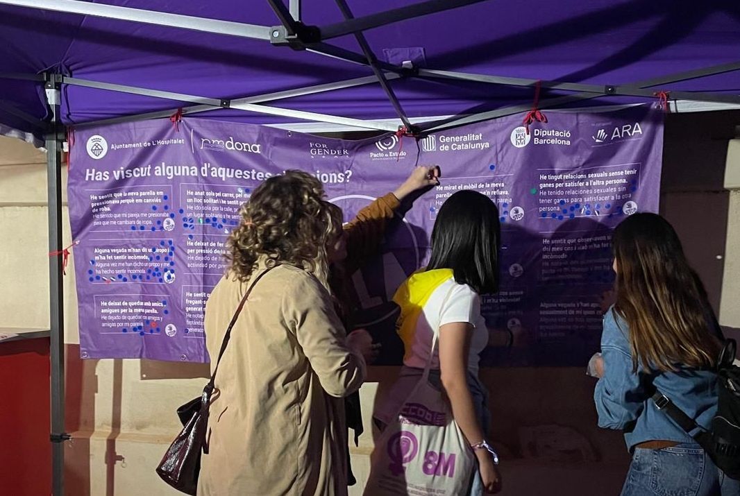 Gent mirant una pancarta lila amb text, sota una tenda.