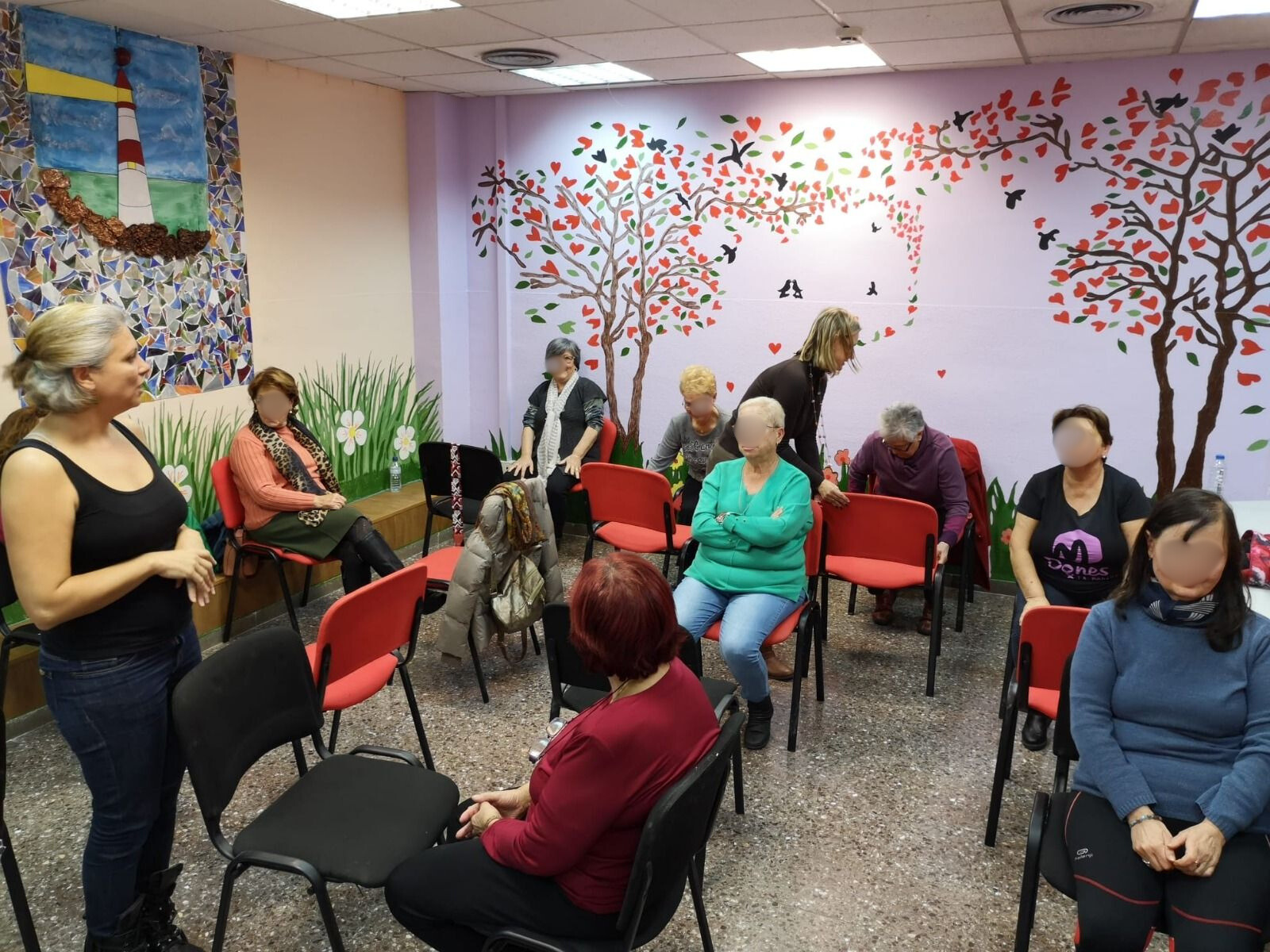 Un grup de persones assegudes en cadires en una sala amb un mural; una dona està dreta i els parla.