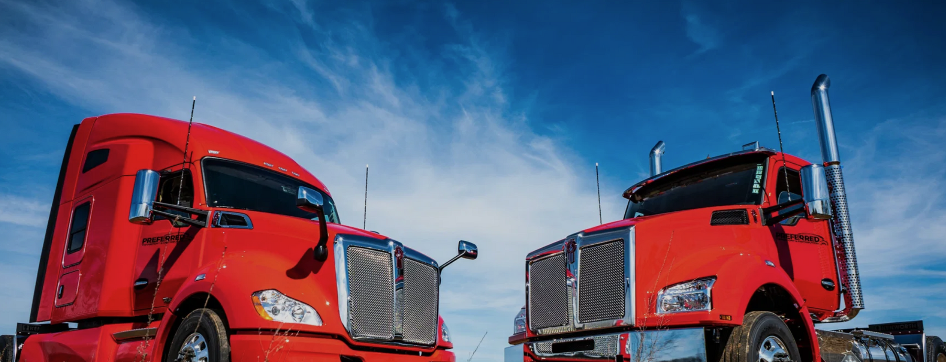 Two red semi-trucks with chrome grills and stacks against a blue sky.