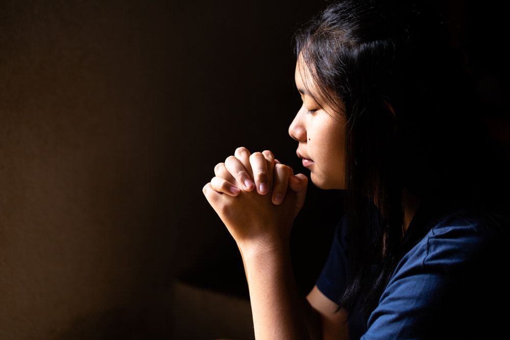 Woman with hands clasped in prayer, eyes closed, against a dark background.