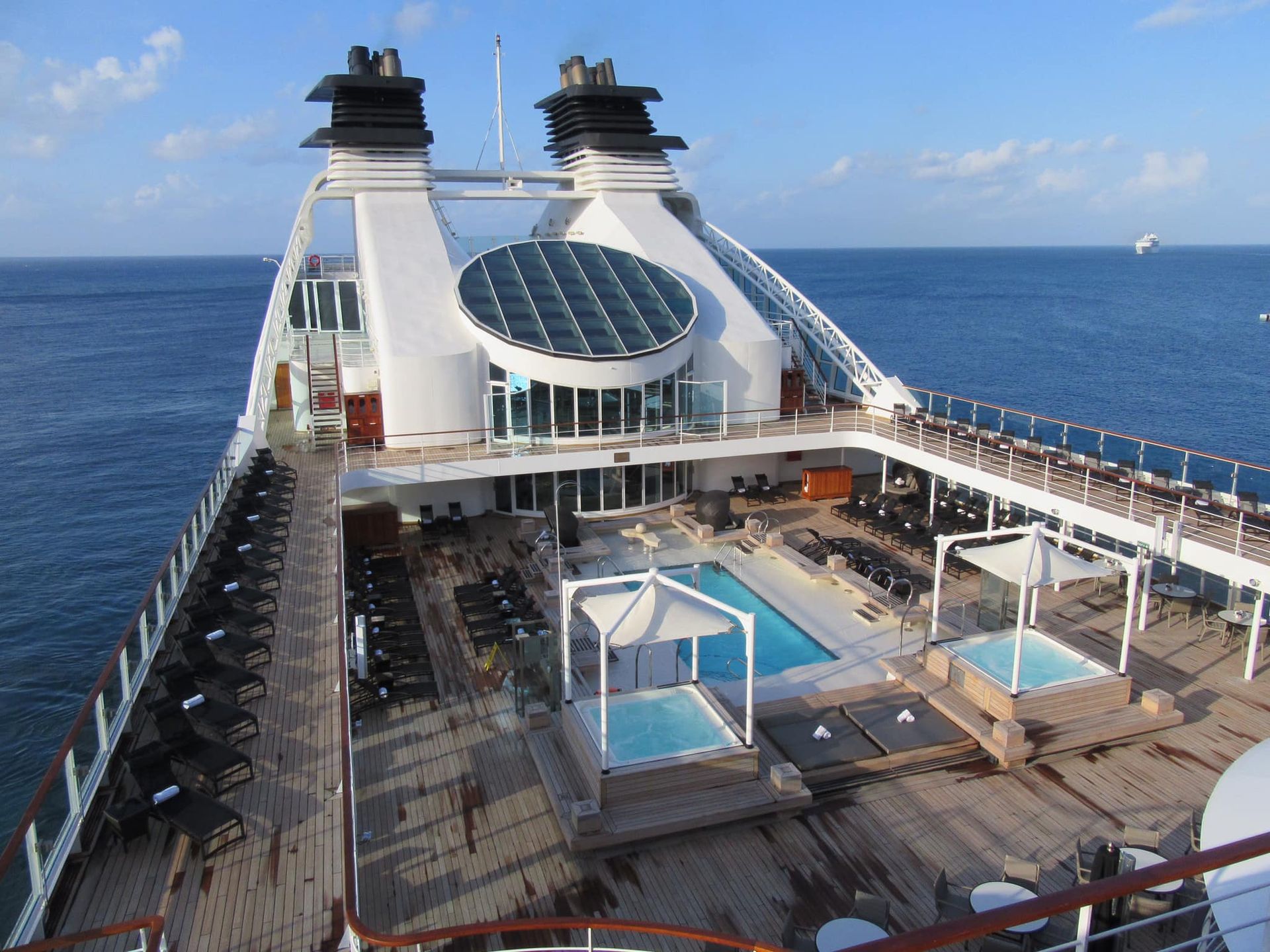 Cruise ship deck with pool, hot tubs, lounge chairs, and ocean view.