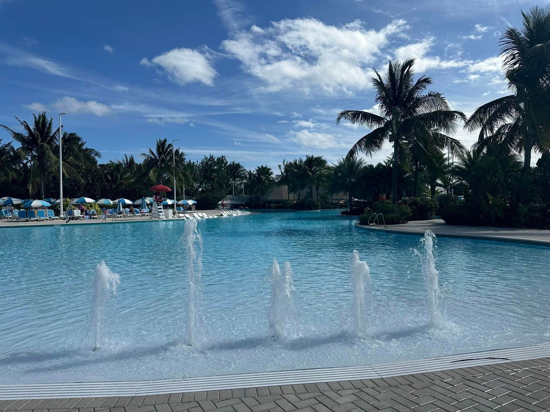 Large blue pool with fountains, palm trees, and a partly cloudy sky.