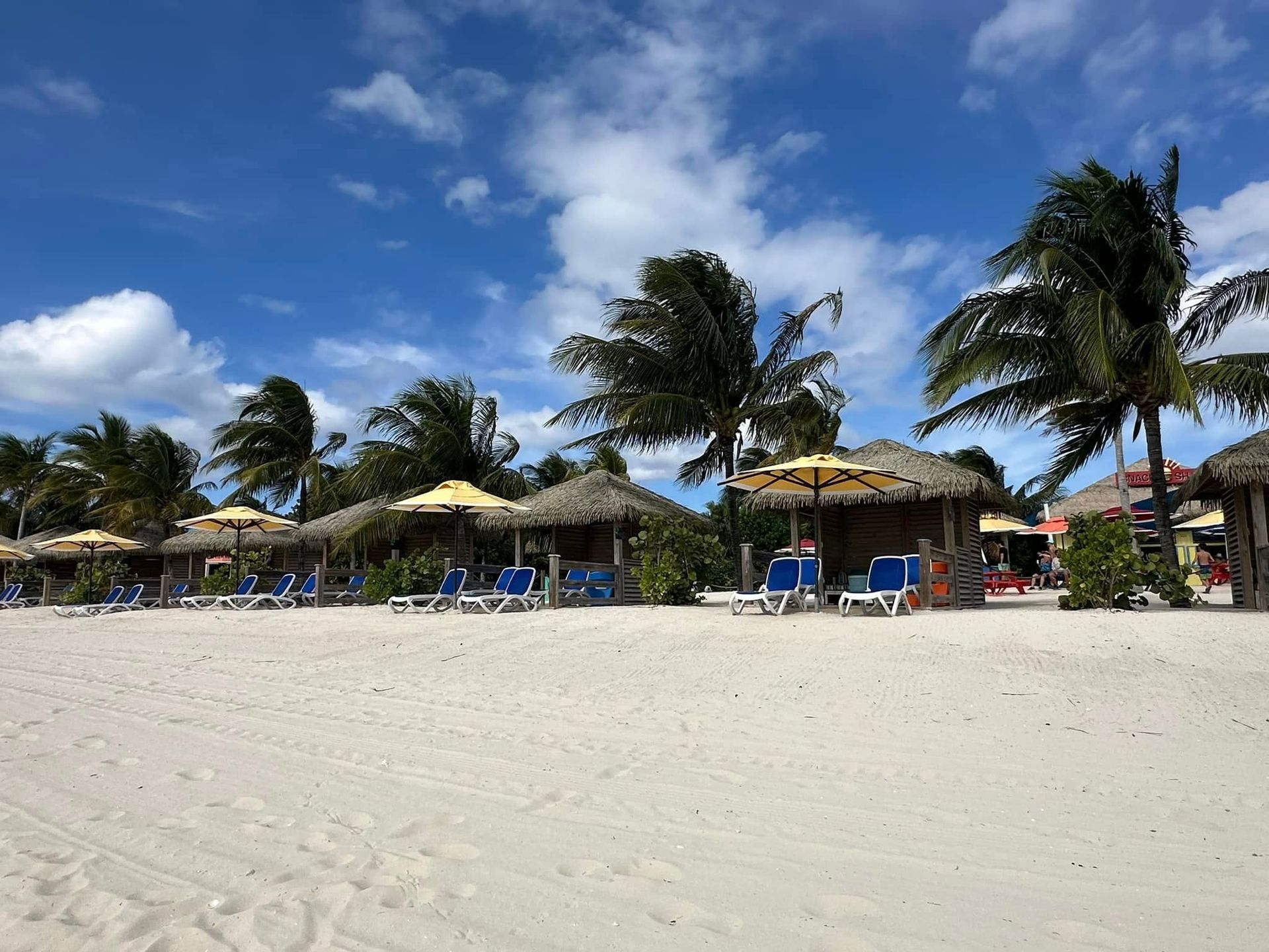 Beach with white sand, palm trees, thatched huts, and lounge chairs under a blue sky with clouds.