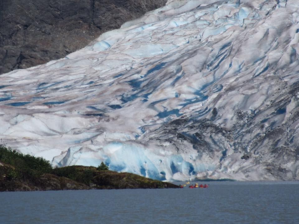 Glacier meeting water; kayaks in foreground. Blue ice with white streaks. Mountain backdrop.