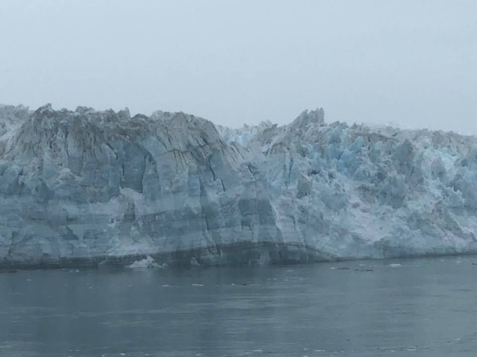 A large, icy blue glacier meets the sea under a grey, overcast sky.