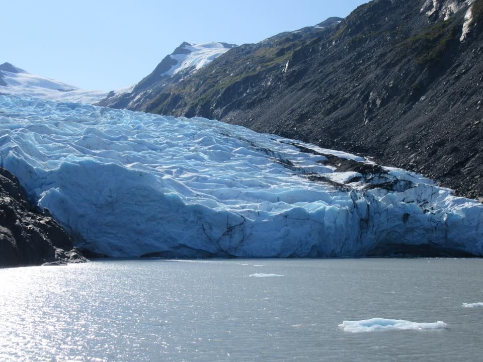 Glacier with blue ice cascading into a lake, flanked by dark mountains under a clear sky.