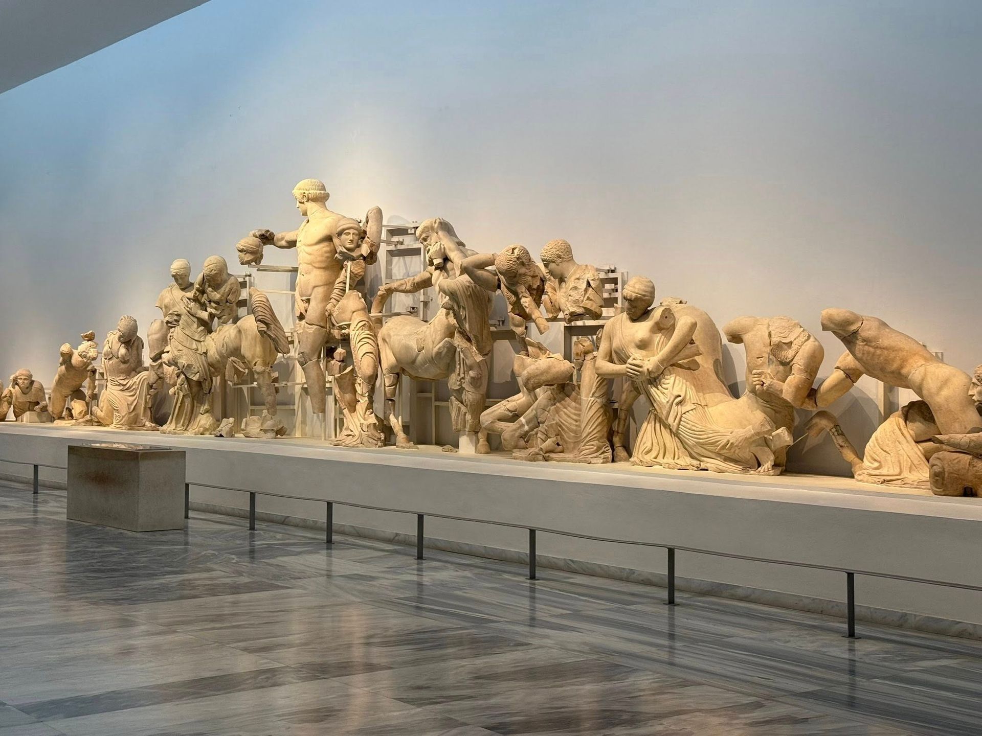 Sculptures from the Parthenon in a museum. Figures in white stone, lined up on a display shelf, lit by overhead lights.
