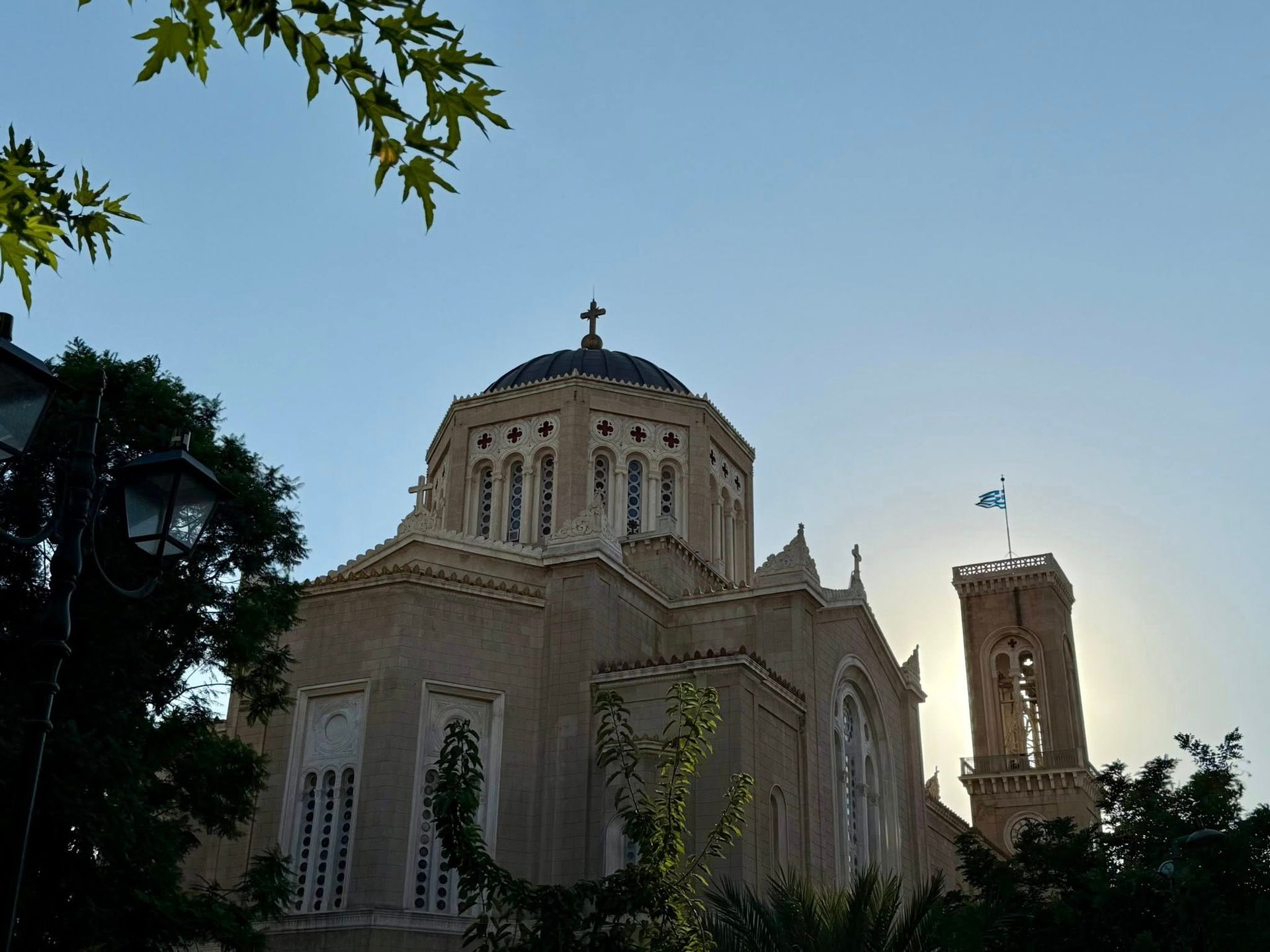 Church with dome and bell tower against a blue sky, Greek flag visible, sun in the background.