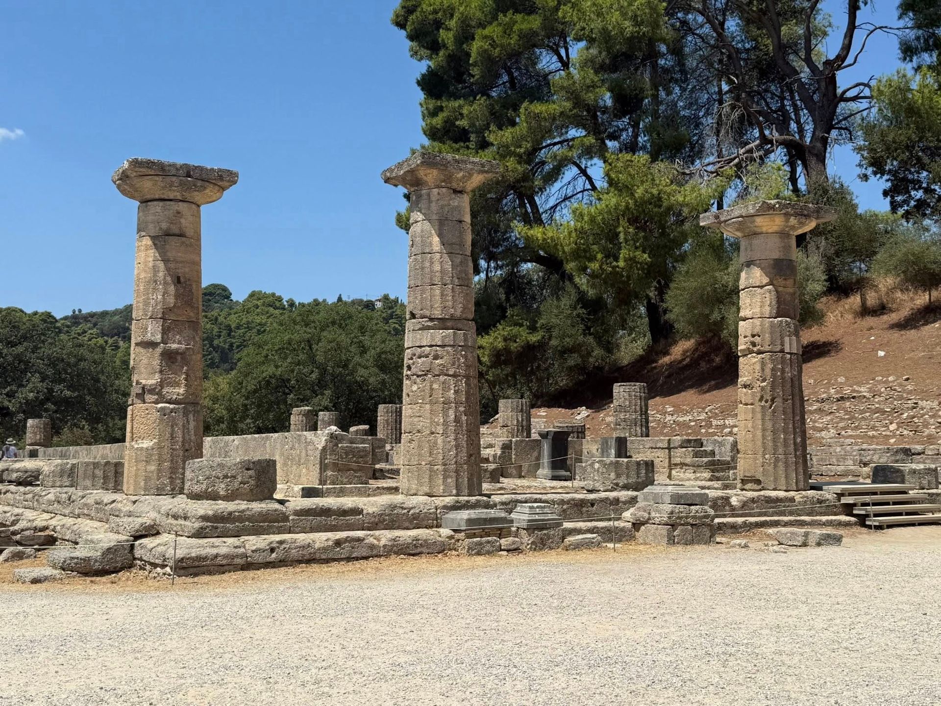 Ruins of an ancient Greek temple with three tall columns in Olympia, Greece, against a backdrop of trees and a blue sky.