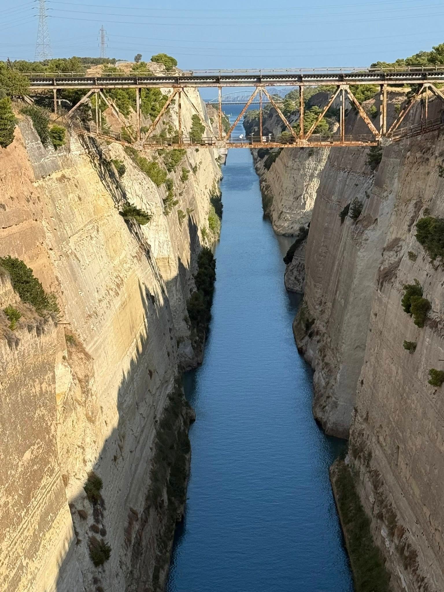 A deep, narrow canal with blue water, flanked by high, light-colored rock walls, crossed by a rusty metal bridge.