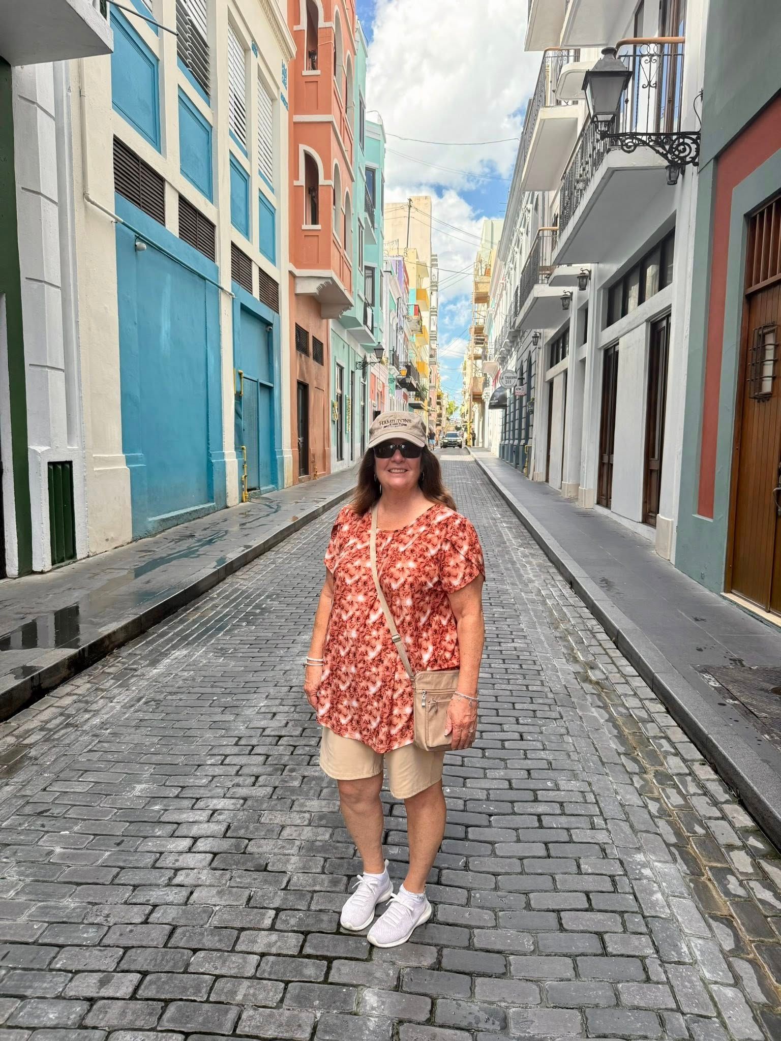 Woman stands on a cobblestone street in Old San Juan, Puerto Rico. She wears a floral shirt, shorts, and a hat. 