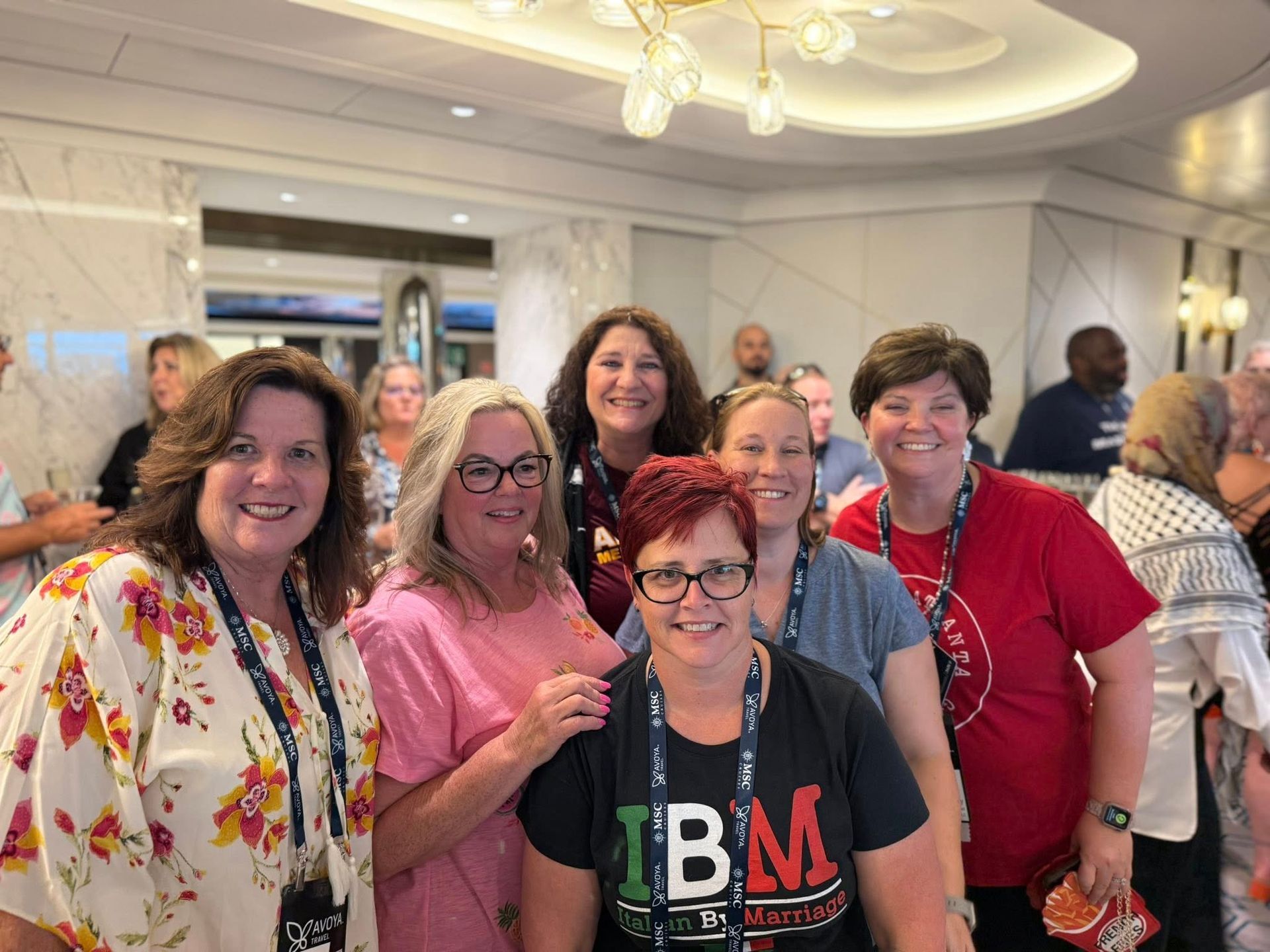 Group of women smiling, standing closely together indoors, some wearing lanyards, warm lighting.