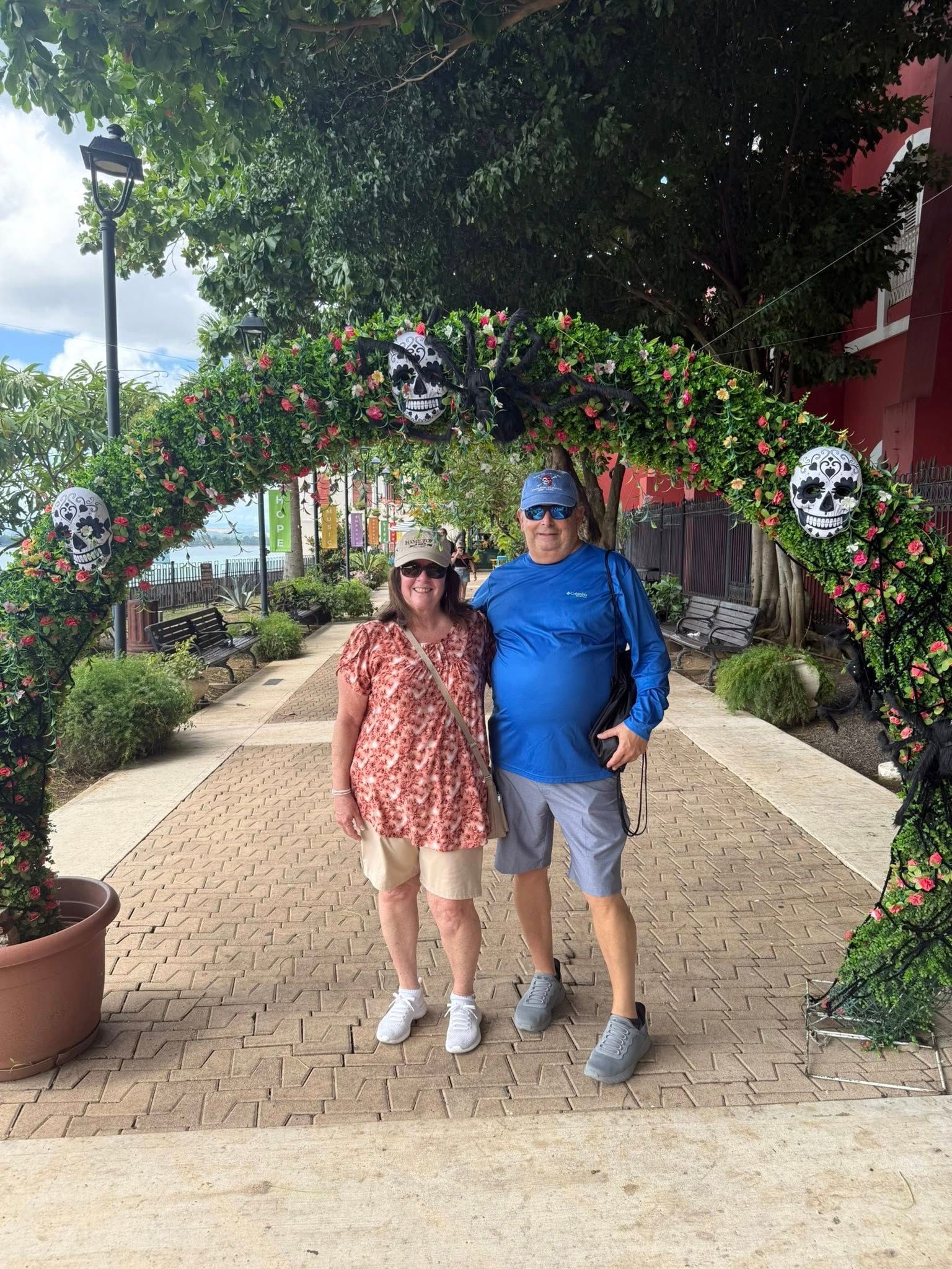 Couple poses under a floral archway with skull decorations, on a brick path.