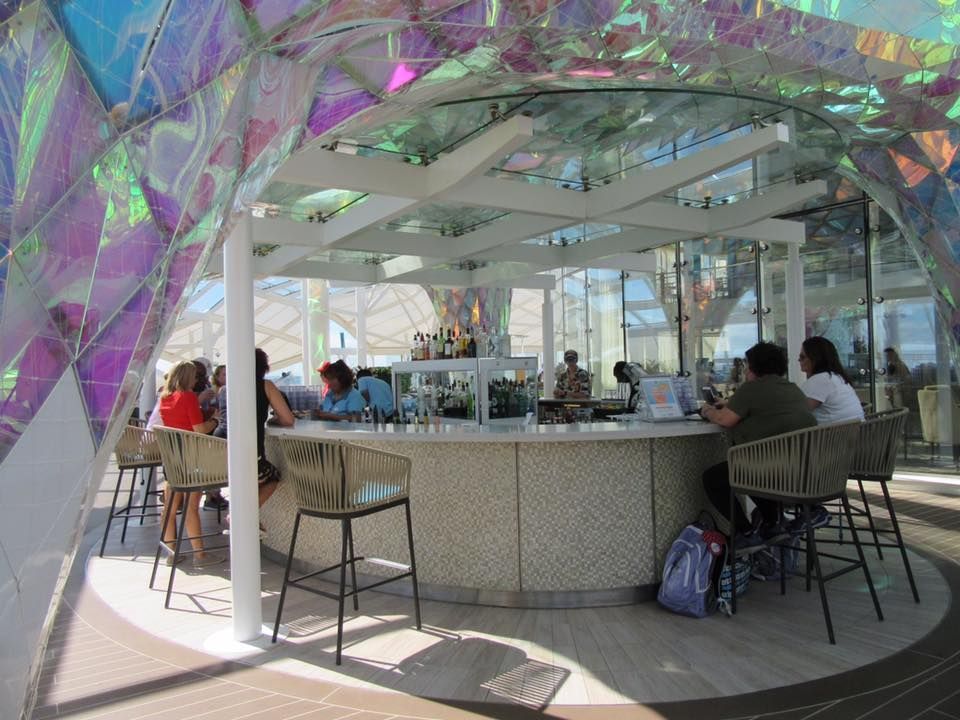 Bar with people seated; iridescent roof, white bar with stools, sunny outdoor setting.