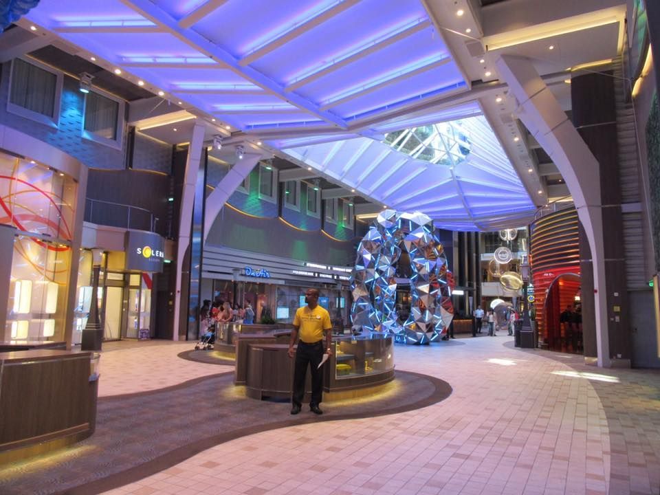 Interior of a brightly lit cruise ship promenade with shops, glass sculptures, and a crew member.