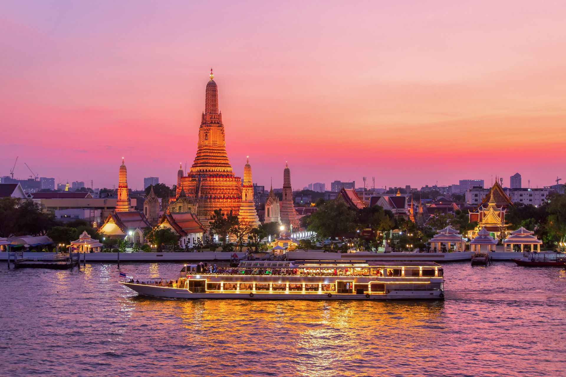 A lit boat sails on a river at dusk, with Wat Arun temple in Bangkok, Thailand illuminated in the background.