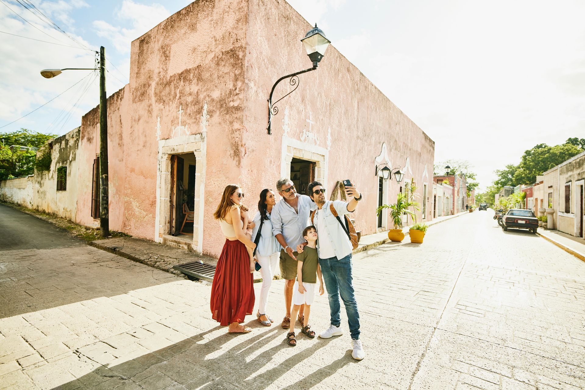 Family standing on a cobblestone street, looking at a pale pink building. Sunlight casts long shadows.