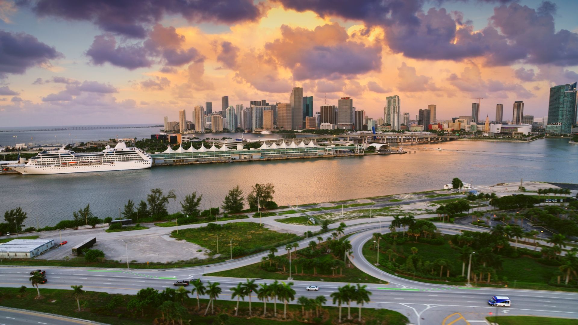 A cruise ship sails through a harbor with city skyline in the background, blue water and sky at dusk.