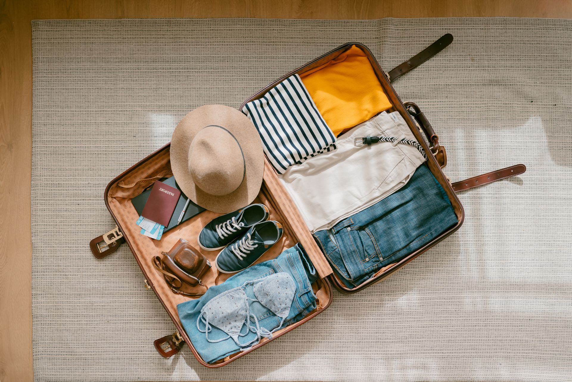 Opened suitcase with clothes, hat, shoes, and passport, on a rug.
