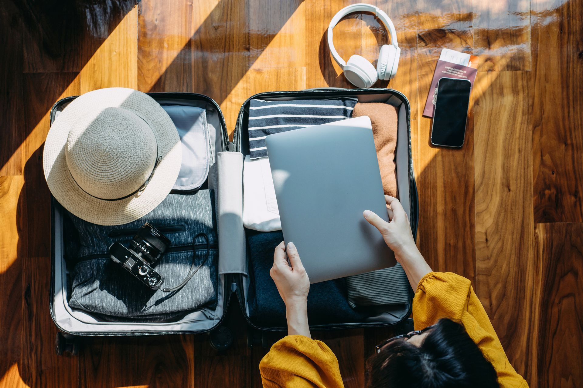 Person packing suitcase with clothes, laptop, hat, headphones, phone, and tickets on a wood floor.