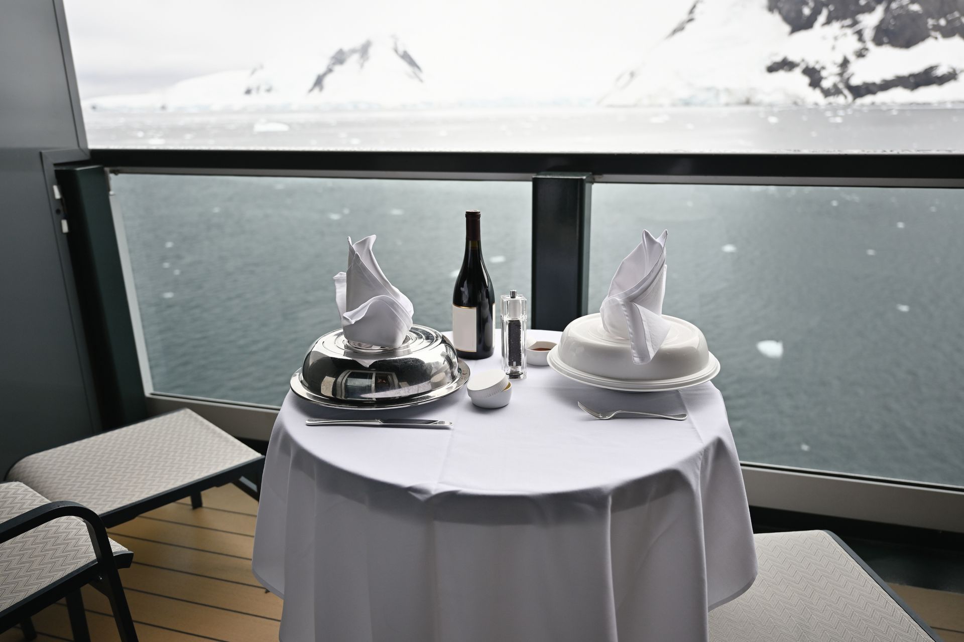 Table set for a meal on a balcony overlooking snowy mountains and water. Silverware, wine, and food under a covered dish.