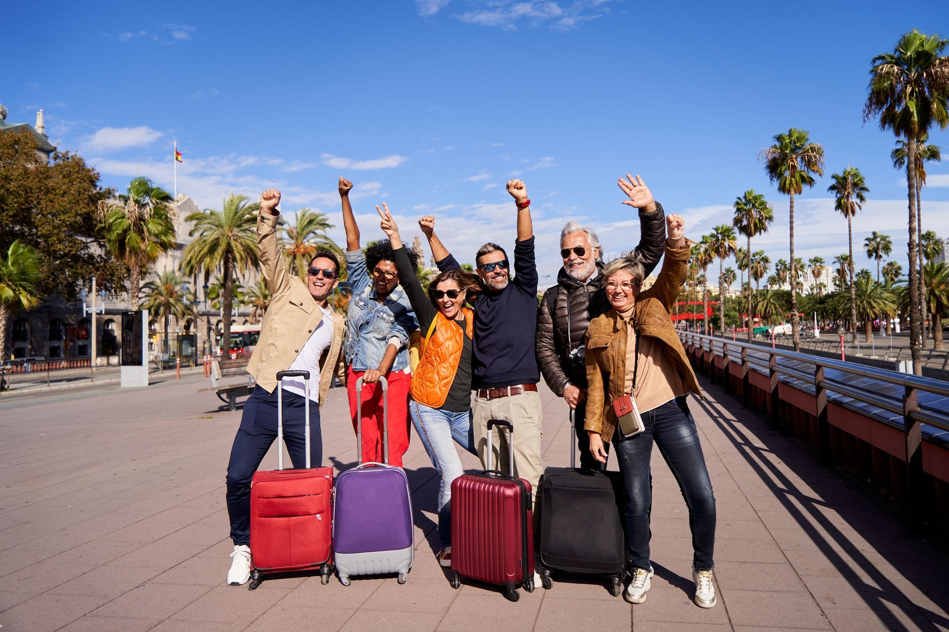 Group of people with luggage, cheering on a sunny boardwalk with palm trees.