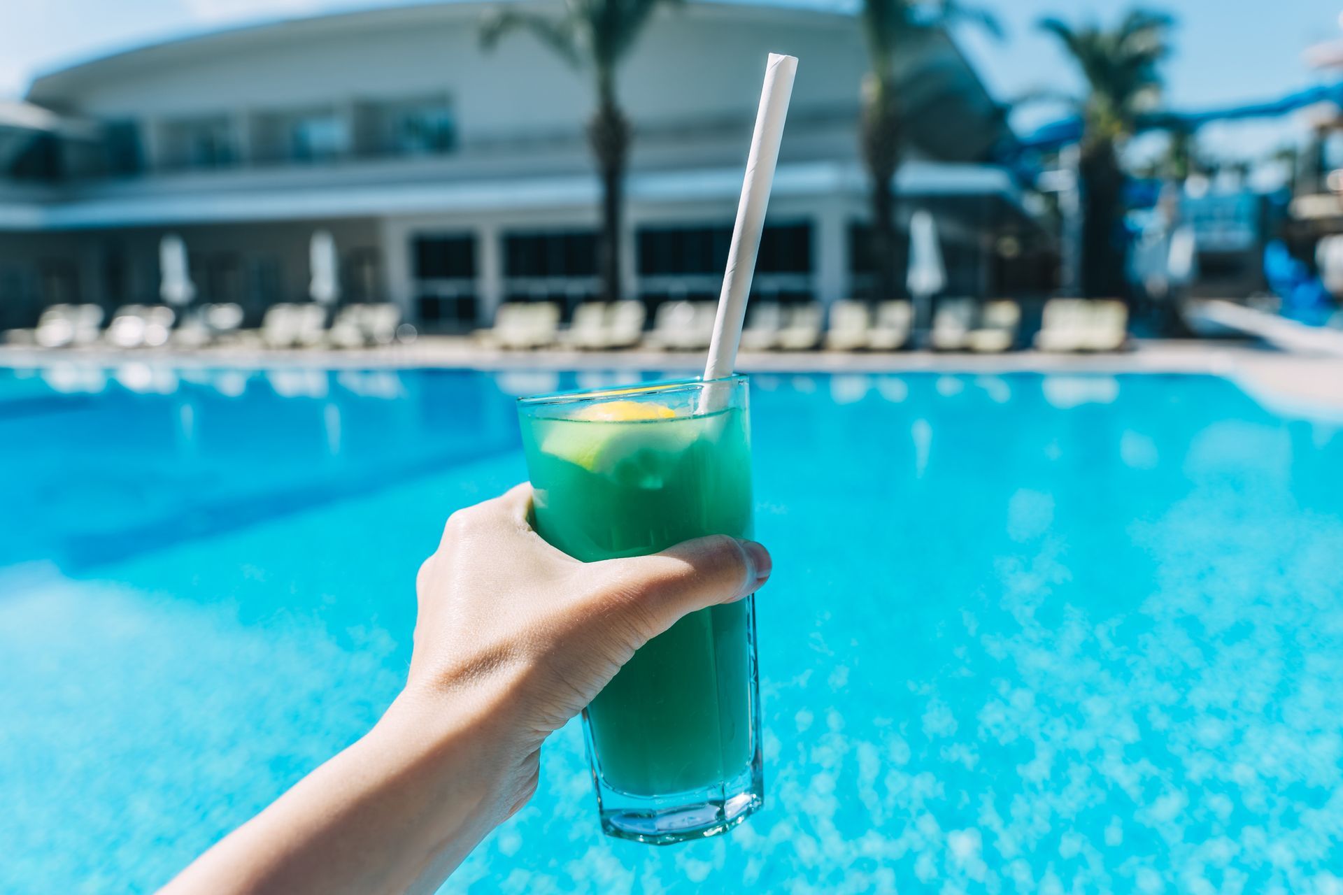 Hand holding a blue cocktail with a straw by a swimming pool.