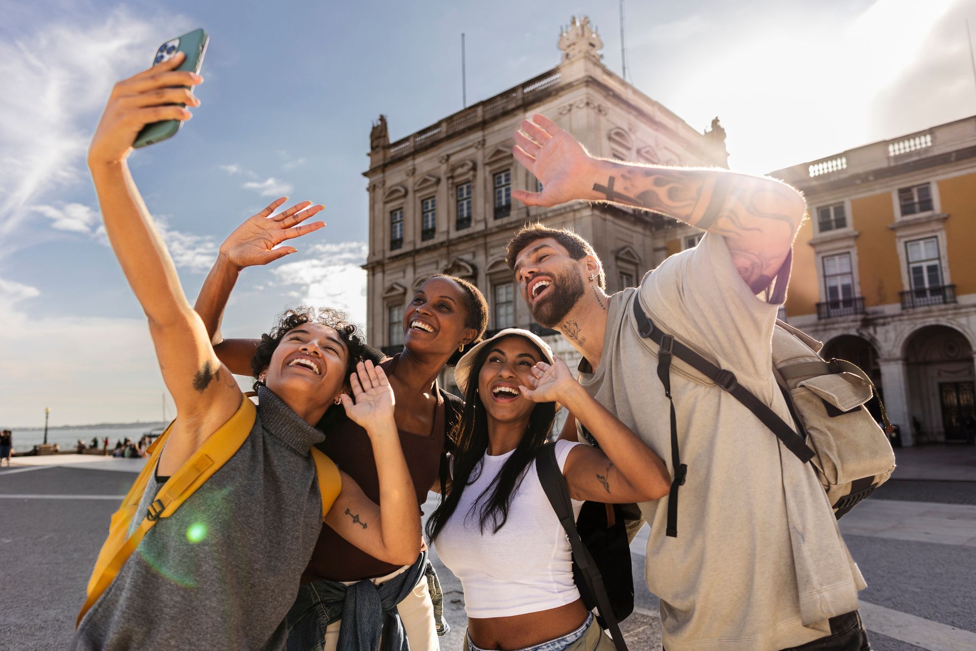 Group of friends taking a selfie, waving, and smiling in front of a building with columns. Bright day.