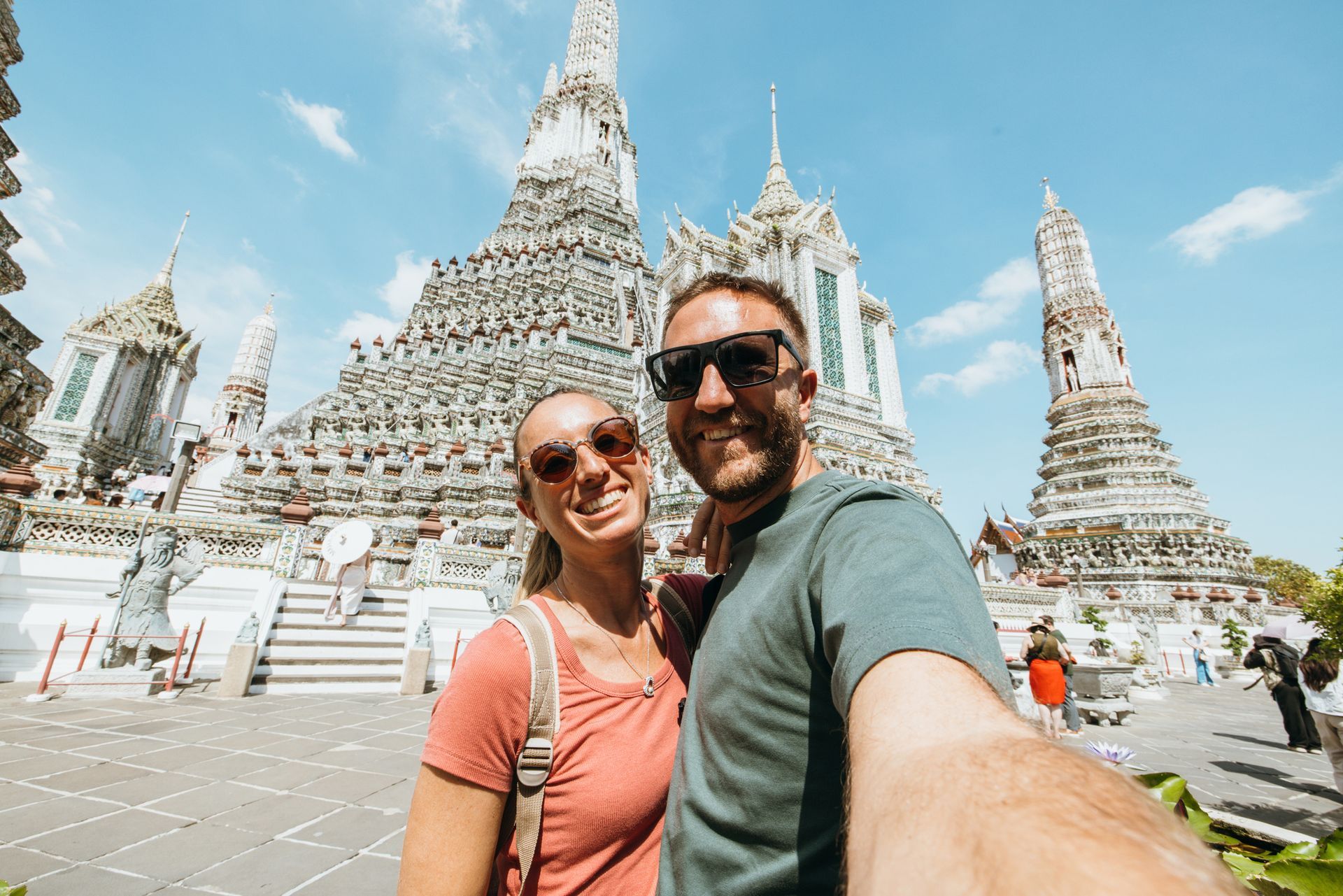 Couple smiles for selfie in front of the ornate white spires of Wat Arun temple in Bangkok, Thailand.