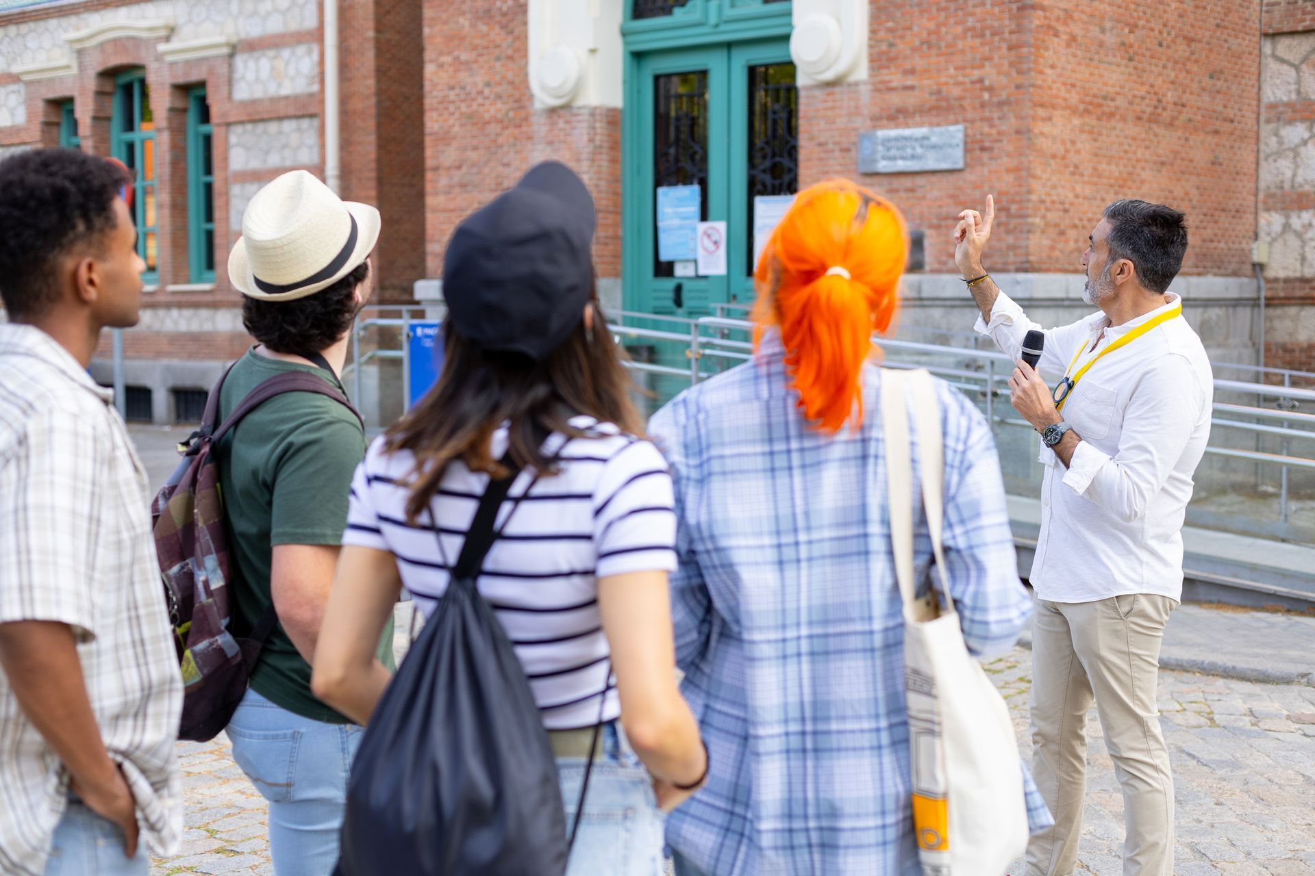 Group of people on a tour, listening to a guide in front of a brick building with green doors.