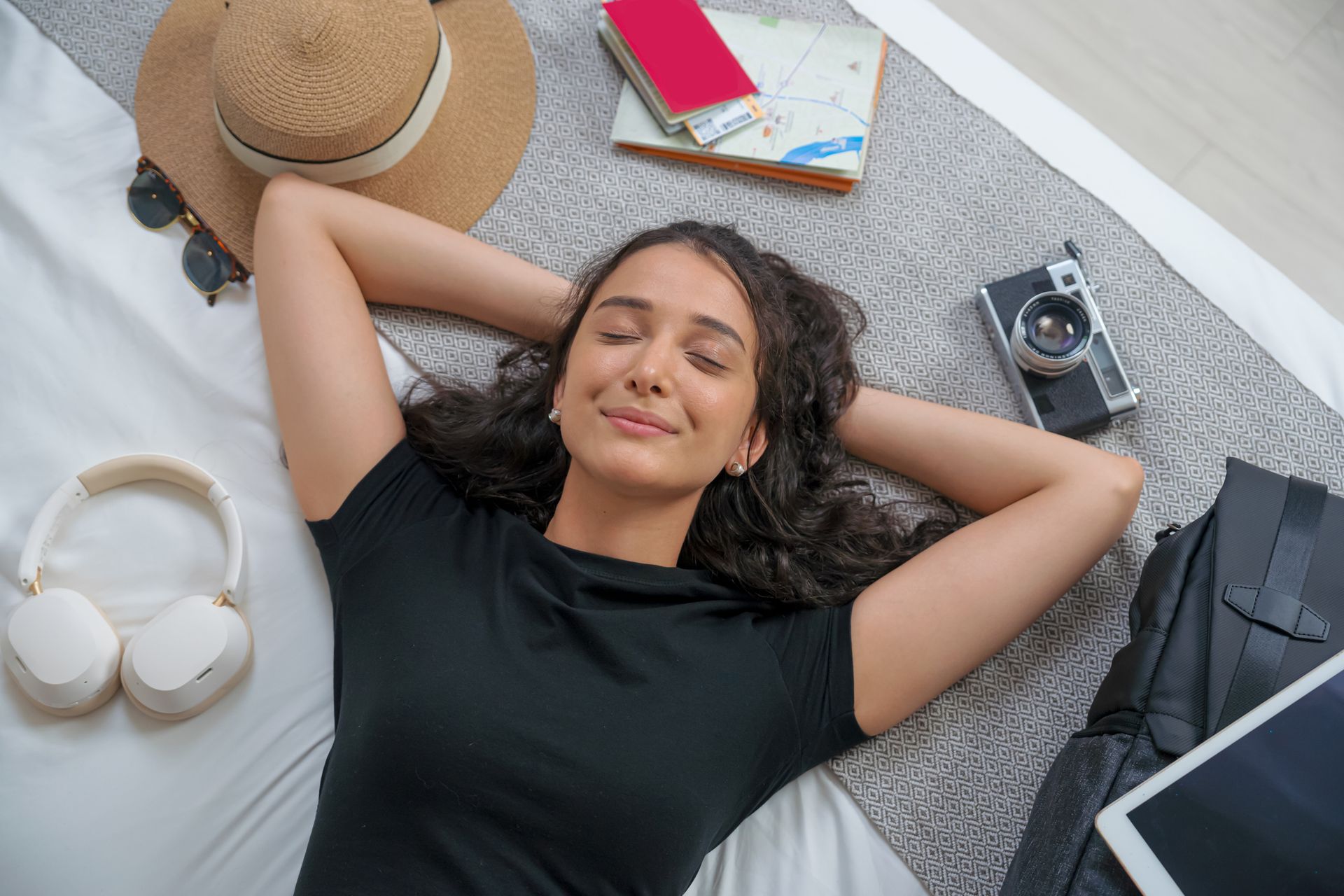 Woman relaxing on bed, surrounded by travel items: hat, headphones, camera, backpack.