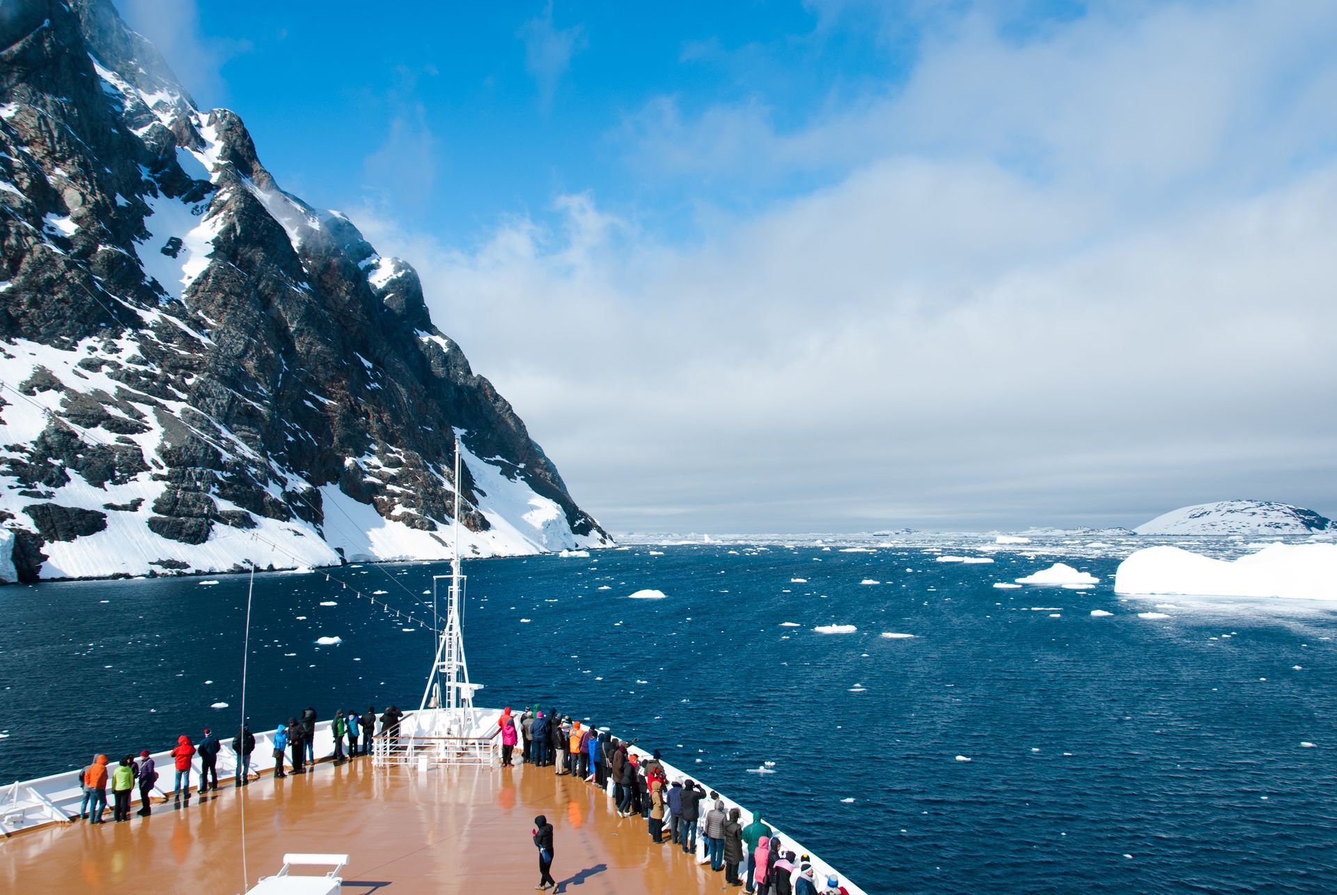 Cruise ship deck with passengers viewing snowy mountains and ice-filled water.