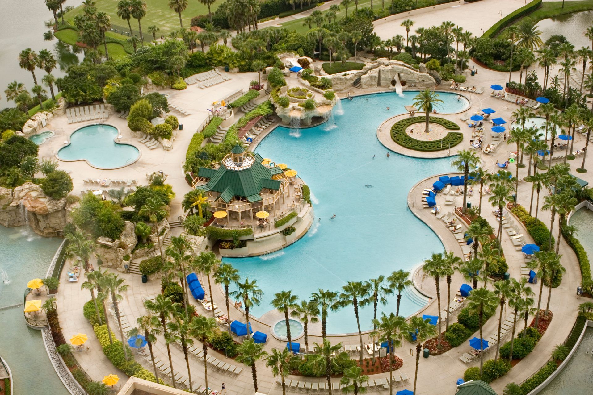 Aerial view of a large, lagoon-style pool area with palm trees, waterfalls, and a building with a green roof.